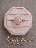 A rose gold ring with an oval pink stone, displayed on a light pink octagonal velvet ring box over a textured gray background, with the brand name 'Angels Blush' written in rose gold script at the bottom.