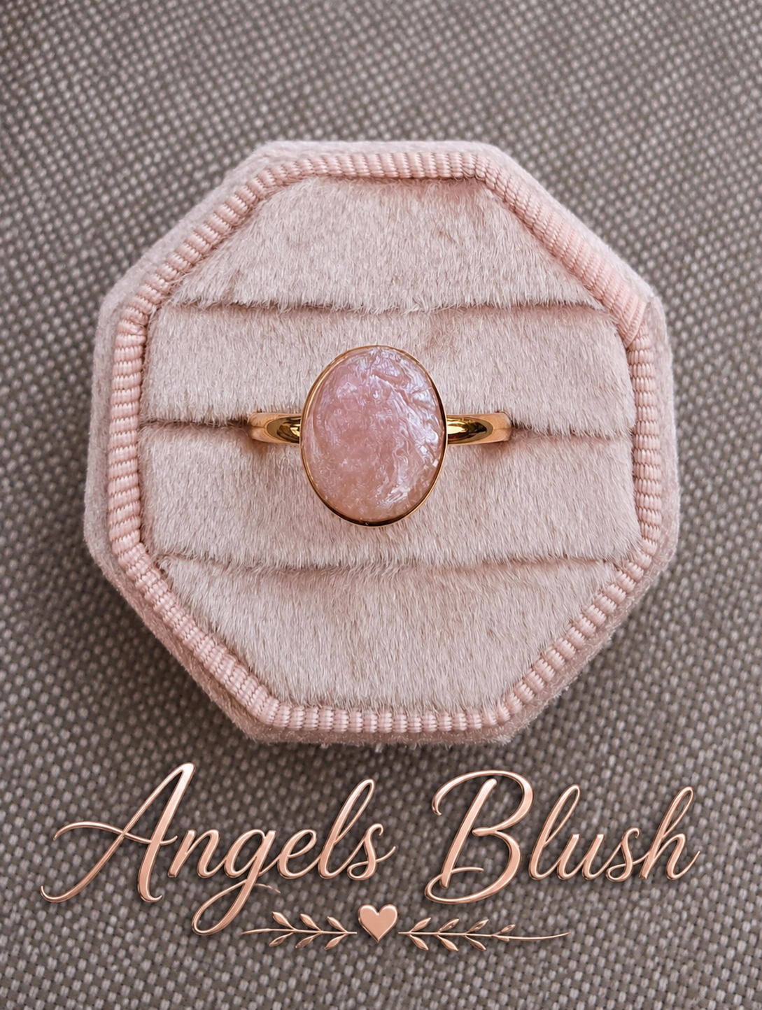 A rose gold ring with an oval pink stone, displayed on a light pink octagonal velvet ring box over a textured gray background, with the brand name 'Angels Blush' written in rose gold script at the bottom.