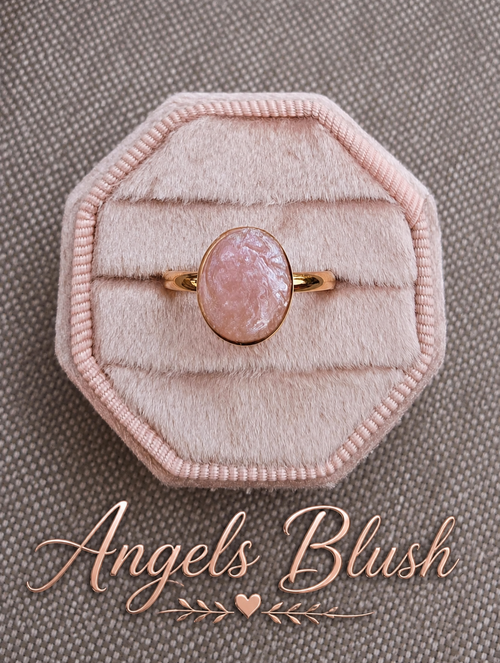 A rose gold ring with an oval pink stone, displayed on a light pink octagonal velvet ring box over a textured gray background, with the brand name 'Angels Blush' written in rose gold script at the bottom.