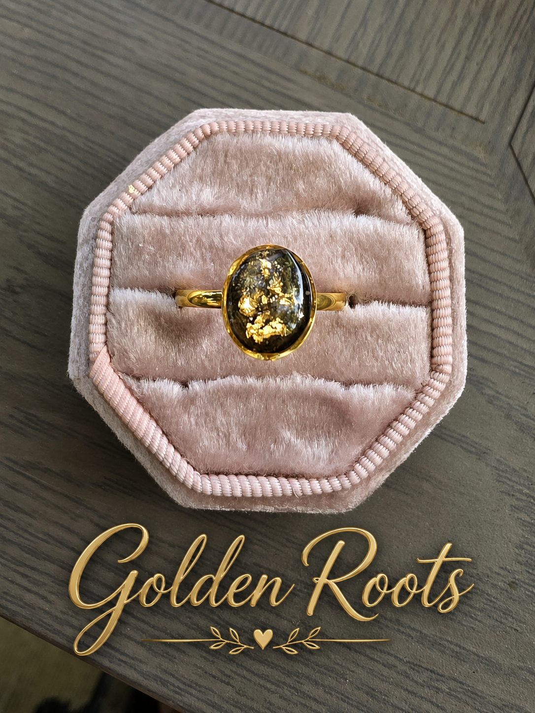 A gold ring with a large oval dark amber or honey-colored stone, displayed in an octagonal pink velvet ring box on a gray wood-grain surface. The brand name 'Golden Roots' is written in gold script at the bottom.