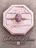 A gold ring with an oval purple gemstone displayed on a pink octagonal velvet ring box, set against a textured gray background with the brand name 'Cherished Bloom' written at the bottom.