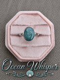 A silver ring with an oval teal-blue stone displayed on a pink velvet ring box, set against a textured gray background with the text 'Ocean Whisper' at the bottom.