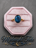 A gold ring with an oval blue stone featuring black marbling, displayed on a pink octagonal velvet ring box. The text 'Midnight Tide' with decorative gold and blue flourishes is written at the bottom.
