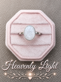 A silver band ring with a round, milky white stone set in the center of a light pink octagonal velvet ring box, placed on a textured gray fabric background with 'Heavenly Light' text and sparkles at the bottom.