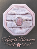 A ring with an oval pink gemstone containing pressed flowers, displayed on a pink octagonal velvet ring box. The text 'Angels Blossom' is written below with a small flower graphic.