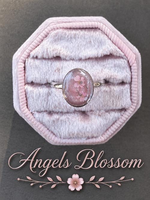 A ring with an oval pink gemstone containing pressed flowers, displayed on a pink octagonal velvet ring box. The text 'Angels Blossom' is written below with a small flower graphic.